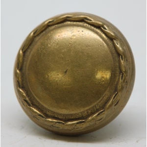 May include: A round, antique brass door knob. The knob has a circular design with a raised, textured border. The center of the knob is smooth and slightly concave. The brass has a warm, aged patina.