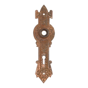 Może przedstawiać: Bogato zdobiona antyczna płytka do klamki drzwiowej z misternymi wzorami kwiatowymi i geometrycznymi. Płytka wykonana jest z metalu i ma miedziany kolor.