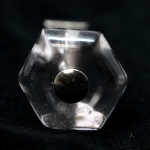 Pode incluir: Um tampão de garrafa hexagonal de vidro transparente com uma tampa de metal prateado. O tampão tem uma aparência nublada e translúcida.