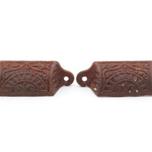 May include: Two antique, cast iron drawer pulls with a rustic, weathered appearance. Each pull features an ornate, symmetrical design with a sunburst pattern and floral accents. The pulls have a dark brown, almost reddish-brown color.