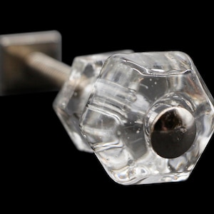 Puede incluir: Un pomo de puerta de cristal transparente con forma hexagonal y un tornillo de metal plateado. El pomo tiene una superficie texturizada en relieve.