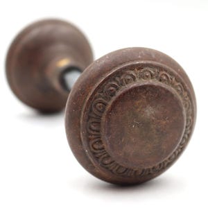 Puede incluir: Primer plano de dos pomos de puerta antiguos de bronce. El pomo delantero está enfocado, mostrando un diseño circular con un patrón ornamental y repetitivo en el borde. Los pomos son de color marrón cálido, sobre un fondo blanco.