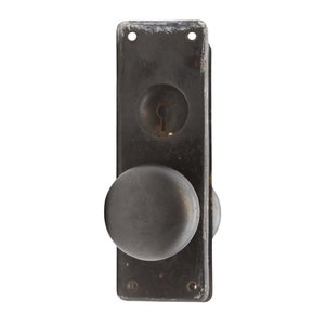 May include: A black metal door knob with a round handle and a keyhole. The knob is attached to a rectangular plate with two screws.