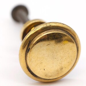 May include: A gold-toned door knob with a round, flat top and a decorative rim. The knob is attached to a metal shaft.