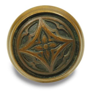 May include: A round, antique-style brass knob with a detailed, symmetrical floral design. The knob has a dark, aged patina, and the floral pattern is raised, creating a textured surface. The knob is likely for a cabinet or drawer.