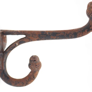 May include: A wall-mounted metal coat hook featuring a double-pronged design with a dark, rusted finish. The hook has a curved upper arm and a smaller lower arm, both ending in rounded finials, with a flat circular base plate for mounting.