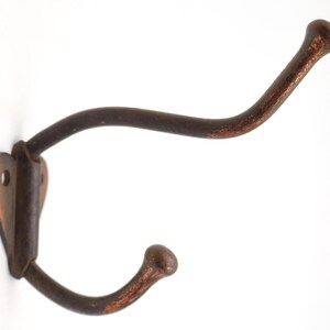 Puede incluir: Un perchero de pared rústico de doble gancho. El gancho de metal tiene un acabado marrón oscuro envejecido con detalles de color óxido. La placa de montaje tiene forma de corazón. Los ganchos se curvan hacia arriba y hacia abajo.