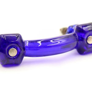 Op de afbeelding: Een levendige blauwe glazen kastgreep met een gebogen handvat en zeshoekige uiteinden. Elk uiteinde heeft een zilverkleurige metalen accent. De hardware is ontworpen voor meubels en voegt een vleugje kleur toe.