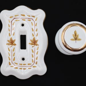 May include: White ceramic light switch cover and matching knob with gold trim and leaf design. The switch plate has a decorative scalloped edge and leaf accents. The knob features a single leaf design.