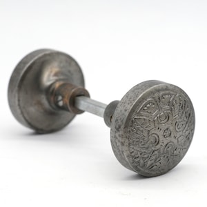 Op de afbeelding: Een vintage zilveren deurklink met een ingewikkeld bloemmotief. De klink is van metaal en heeft een licht verweerde afwerking.