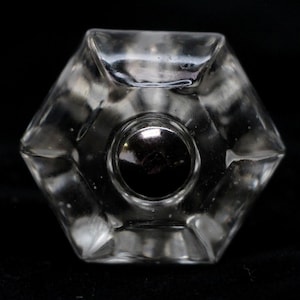 Puede incluir: Un tapón de botella de vidrio transparente con forma hexagonal y una parte superior de metal plateado.