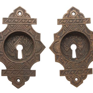Op de afbeelding: Twee antieke messing sleutelgaten met ingewikkelde geometrische ontwerpen. Elk sleutelgat heeft een ronde opening met een sleutelgat in het midden.