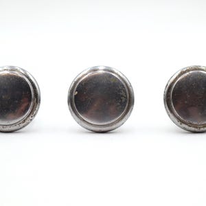 Peut inclure: Trois boutons de meuble ronds et antiques en métal argenté. Chaque bouton a un centre légèrement bombé et un bord décoratif en relief. Les boutons sont disposés horizontalement sur un fond blanc.