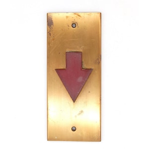 May include: A vintage brass elevator button plate featuring a red downward arrow. The rectangular plate, with two screw holes, has a distressed, aged appearance, indicating it is an antique or reclaimed item.