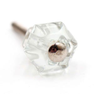 May include: A clear, faceted glass cabinet knob with a silver-colored metal base and screw. The knob has a hexagonal shape and is set against a white background. This decorative hardware is suitable for drawers and cabinets.