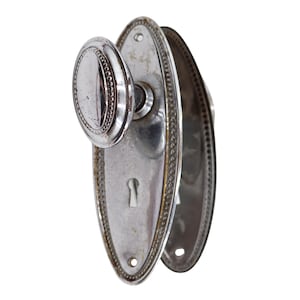 May include: A vintage silver-toned door handle set. The oval-shaped knob and backplates feature a beaded edge design. The keyhole is visible on the backplate. The hardware is likely made of metal and shows signs of wear.
