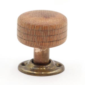May include: A round wooden cabinet knob with a visible grain pattern and three horizontal engraved lines around the side. The knob is attached to a cylindrical metal stem and a circular brass base plate featuring two small screw holes for mounting.