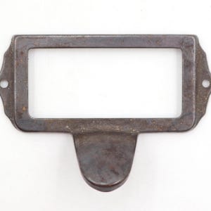May include: A rectangular, antique-style label holder made of dark, weathered metal. The frame has rounded corners and a small, curved handle at the bottom. Two small holes are on either side for mounting. The label area is empty.