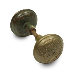 May include: Antique brass doorknob with an ornate, embossed design. The doorknob has a weathered appearance with shades of green and brown, suggesting age and use. The design features swirling patterns and a crown-like element.