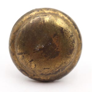 Può includere: Primo piano di un pomello rotondo in ottone antico. Il pomello ha una superficie usurata e strutturata con variazioni di tonalità oro e marrone. L'hardware è probabilmente per un armadio o un cassetto.