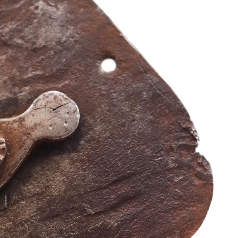 May include: Close-up of a weathered, rusty metal object with a small, circular hole and a riveted, oval-shaped metal piece. The object has a rough, textured surface and appears aged, possibly antique.