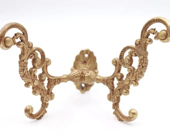Può includere: Gancio da parete ornato, in stile antico, con finitura dorata. Il gancio presenta un design simmetrico con dettagli floreali a volute e estremità curve. La piastra di montaggio centrale ha un design a foglia decorativo. Un pezzo di ispirazione vintage.