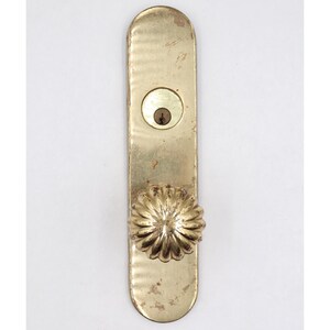 Op de afbeelding: Een vintage messing deurklink met een sleutelgat en een decoratieve, bloemvormige knop. De klink is rechthoekig met afgeronde randen en een getextureerd oppervlak. Het messing heeft een licht verouderd uiterlijk.