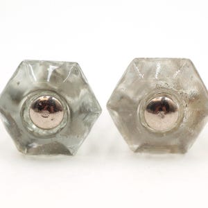 May include: Two clear, hexagonal glass cabinet knobs with silver-colored metal centers. The knobs are vintage and have a slightly aged appearance, suggesting they are antique or reclaimed. They are set against a white background.