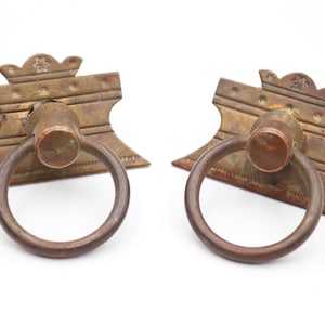 May include: Two antique brass drawer pulls with ring handles. Each pull features a rectangular backplate with decorative details and a circular ring handle. The brass has a warm, aged patina, suggesting vintage craftsmanship.