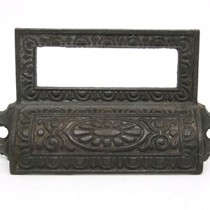 Puede incluir: Tirador de cajón de hierro fundido negro con un diseño ornamentado. El tirador tiene una abertura rectangular para una etiqueta.