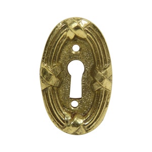 May include: A gold oval-shaped keyhole cover with an intricate design. The cover has a keyhole in the center and a decorative pattern around the edges.