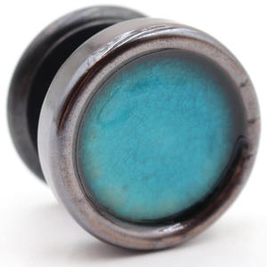 May include: A dark brown wooden ear plug with a teal-colored, crackled glass-like center. The plug has a dark metallic rim and a rounded shape, suitable for stretched ear piercings.