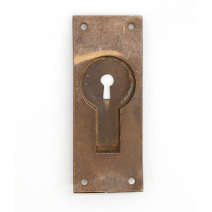 Puede incluir: Una tapa de mirilla de latón con forma rectangular y bordes redondeados. La tapa tiene dos pequeños agujeros en la parte superior e inferior, y un gran agujero de llave en el centro.
