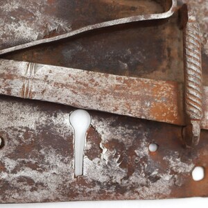 May include: Close-up of a weathered, antique metal lock with a keyhole. The lock has a rectangular base with a sliding bolt and a decorative, twisted handle. The metal shows signs of rust and wear, with a mix of brown and silver tones.