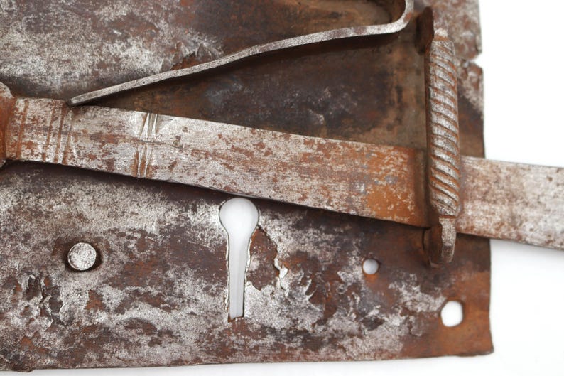 May include: Close-up of a weathered, antique metal lock with a keyhole. The lock has a rectangular base with a sliding bolt and a decorative, twisted handle. The metal shows signs of rust and wear, with a mix of brown and silver tones.