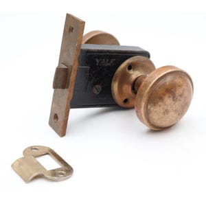Puede incluir: Un juego de cerradura de puerta antigua con acabado de latón. El juego incluye una placa de cerradero rectangular, un cuerpo de cerradura negro con la marca "YALE" y dos pomos redondeados. También hay una placa de ojo de cerradura separada.