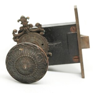 Op de afbeelding: Antiek sierlijk deurknop en slotset met een decoratief bloemmotief. De knop is gemaakt van metaal en heeft een donkerbruine afwerking. Het slot is ook gemaakt van metaal en heeft een zwarte afwerking.