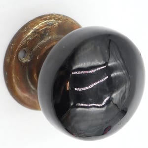 Könnte beinhalten: Ein runder, glänzend schwarzer Türknauf mit einer glatten, reflektierenden Oberfläche. Der Knauf ist an einer kreisförmigen, verwitterten, messingfarbenen Metallplatte befestigt, die Anzeichen von Oxidation und Abnutzung aufweist, inklusive eines sichtbaren Schraublochs.