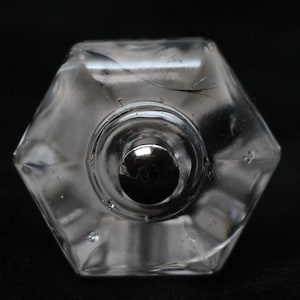 Op de afbeelding: Een glazen flesstopper van helder glas met een zilveren metalen bovenkant. De stopper heeft een zeshoekige vorm en een getextureerd oppervlak.