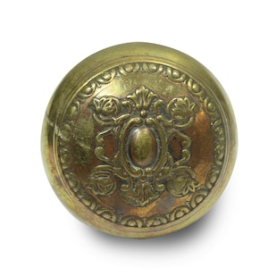 Puede incluir: Un pomo de puerta antiguo de latón con un diseño en relieve ornamentado. El pomo redondo presenta un óvalo central y detalles florales, rodeado por un borde decorativo. El latón tiene una pátina cálida y envejecida.