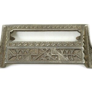 May include: Antique silver drawer pull with an ornate design. The pull has a rectangular opening for a label and two holes for screws.
