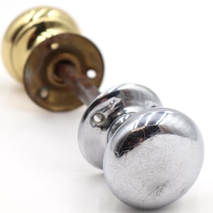 Könnte beinhalten: Zwei antike Türknöpfe. Einer ist goldfarben, der andere silberfarben. Der goldene Türknauf hat eine kreisförmige Basis mit Schraubenlöchern. Der silberne Türknauf ist ein Satz von zwei runden Knöpfen, die an einer Stange befestigt sind.