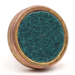May include: A round, decorative cabinet knob with a teal speckled center. The knob has a copper-colored outer ring, a gold-colored middle ring, and a teal center. The knob is designed to be attached to a cabinet or drawer.
