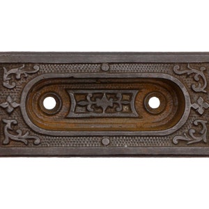 Puede incluir: Una placa de puerta de hierro fundido con una abertura ovalada y detalles ornamentados. La placa es de color gris oscuro con una pátina oxidada.