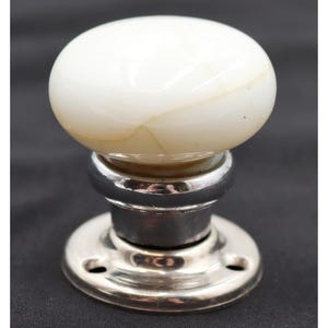 May include: A round, white, marble-like stone door knob with subtle tan veining. The knob is mounted on a polished, silver-toned metal base featuring a circular plate with two visible screw holes for attachment to a door or cabinet surface.
