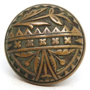 May include: A round, antique bronze button with intricate embossed floral and geometric patterns. The button features a central band of cross-like designs, surrounded by stylized leaves and triangular shapes. The aged finish gives it a vintage look.