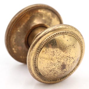 May include: Antique brass door knob with a round, slightly domed shape. The knob has a decorative beaded edge and shows signs of age and wear, with a tarnished finish. The backplate is also round and has a similar beaded edge.