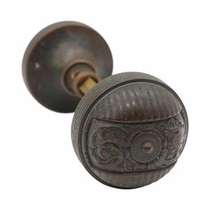 Könnte beinhalten: Antikes Türknauf-Set aus Bronze. Das Set enthält zwei runde Knöpfe, einen mit einem dekorativen Blumendesign und vertikalen Linien und den anderen in einfacher runder Form. Die Knöpfe sind durch eine Messingstange verbunden.