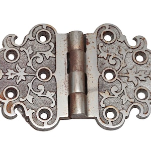 Puede incluir: Una bisagra de metal plateada con diseños florales ornamentados y un acabado desgastado. La bisagra tiene ocho agujeros para tornillos y un pasador de bisagra decorativo, ligeramente oxidado.