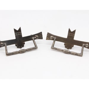 Może przedstawiać: Dwa antyczne srebrne uchwyty do szuflad z ozdobnym wzorem. Każdy uchwyt przedstawia stylizowanego ptaka z koroną na głowie i kwiatowym wzorem u dołu.